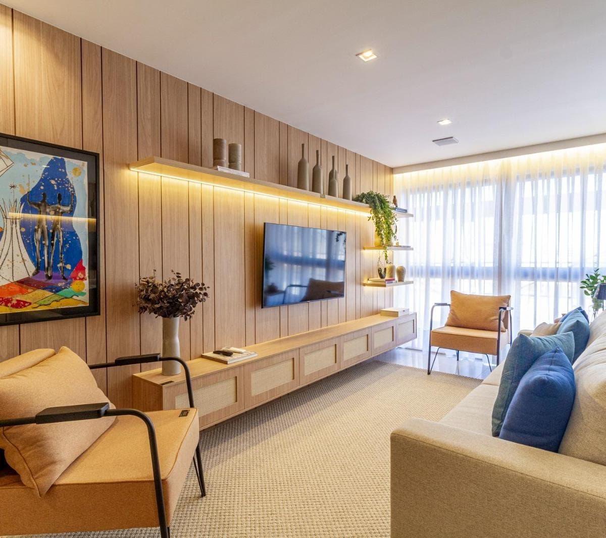 Sala de estar com painel ripado em madeira, TV fixada, sofá cinza e poltronas.