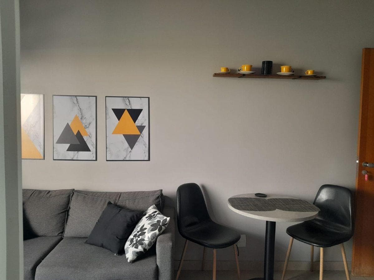 Sala de estar com sofá cinza, mesa redonda com duas cadeiras pretas e quadros decorativos na parede.