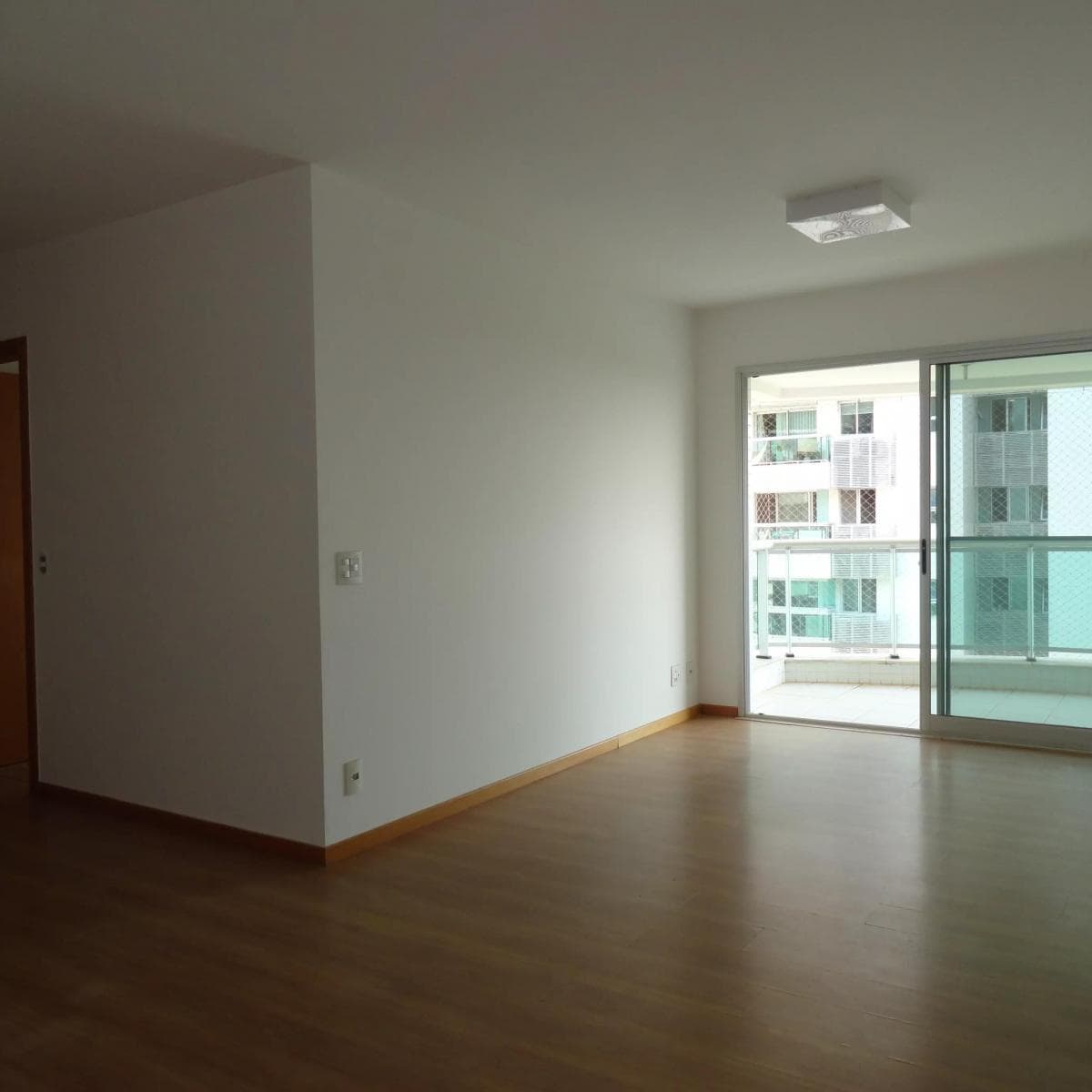 Sala vazia com piso amadeirado, paredes brancas e acesso a varanda.