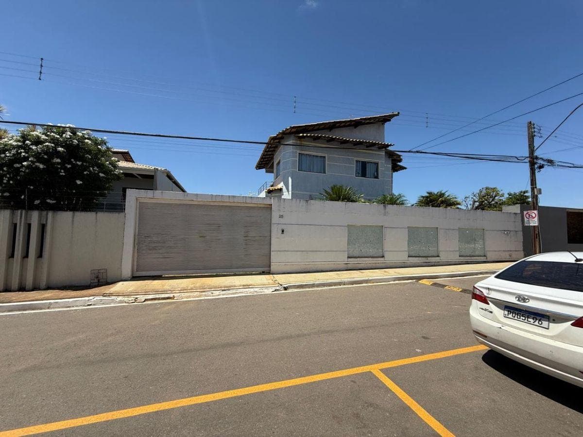 Fachada da casa com muro branco, portão de garagem fechado e sobrado ao fundo.