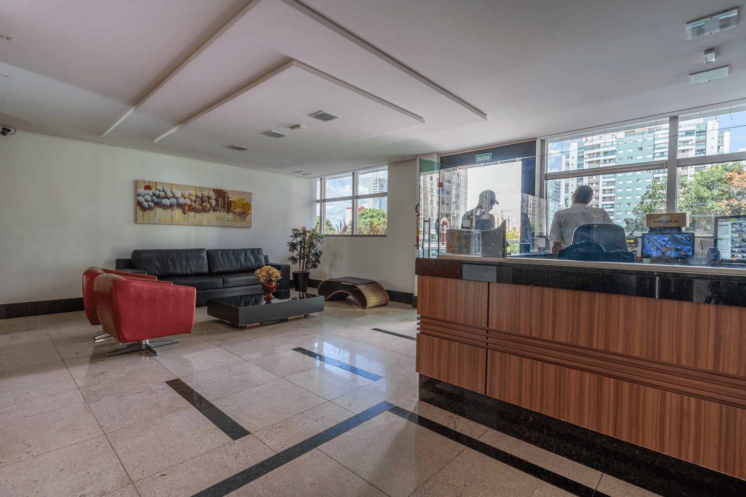 Recepção do condomínio com balcão de atendimento, sofá preto, poltronas vermelhas e piso de mármore.