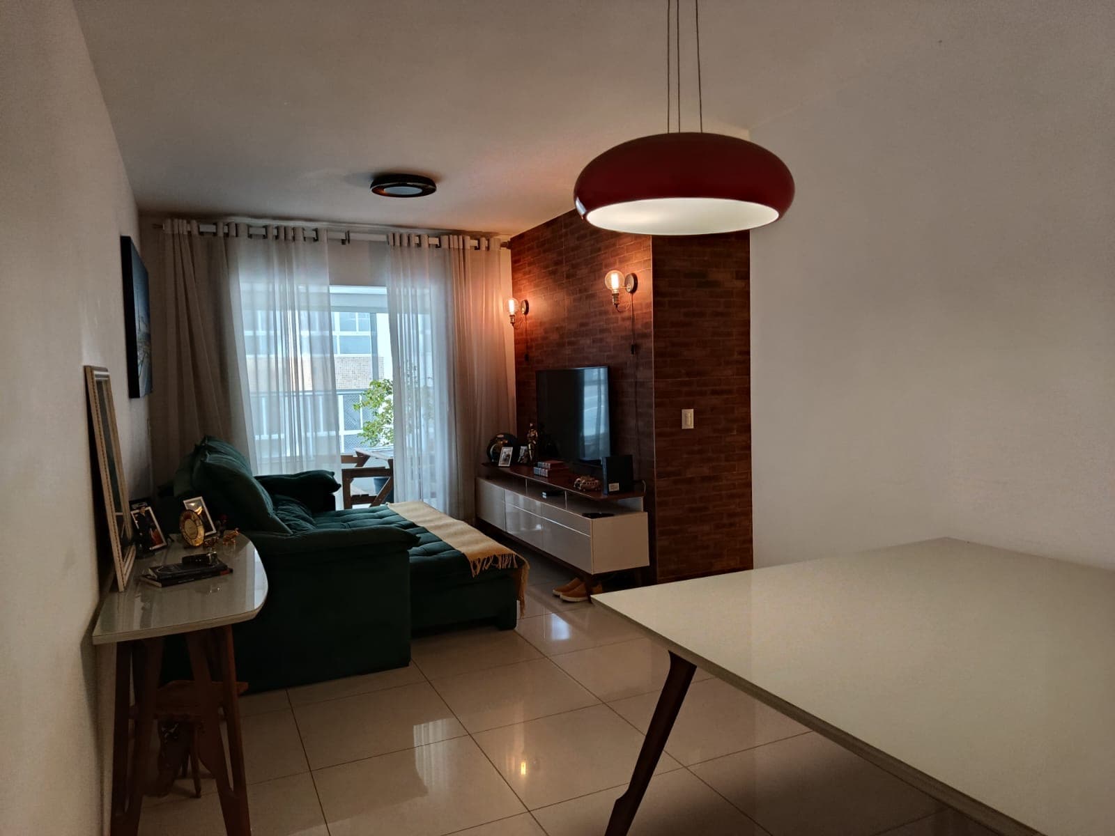 Sala de estar e jantar integrada, com sofá verde, parede de tijolinho à vista e iluminação pendente.